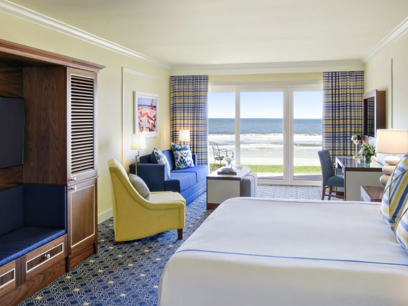 A hotel room featuring a king bed with white linens, and upholstered blue headboard. In the background, a desk and sitting area look out onto the beach beyond.