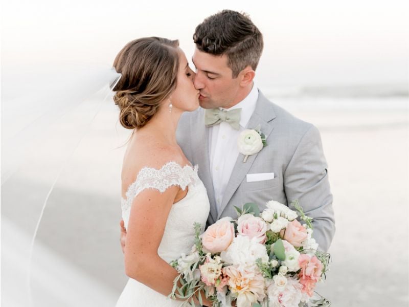 A bride and groom kiss on the beach while holding a bouquet of white and pink flowers.