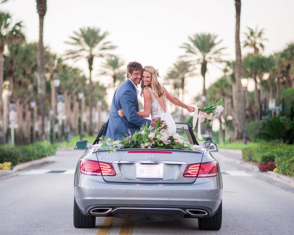 A bride and groom sit on the back of a Mercedes convertible looking behind them and smiling with a palm tree-lined street in the background.