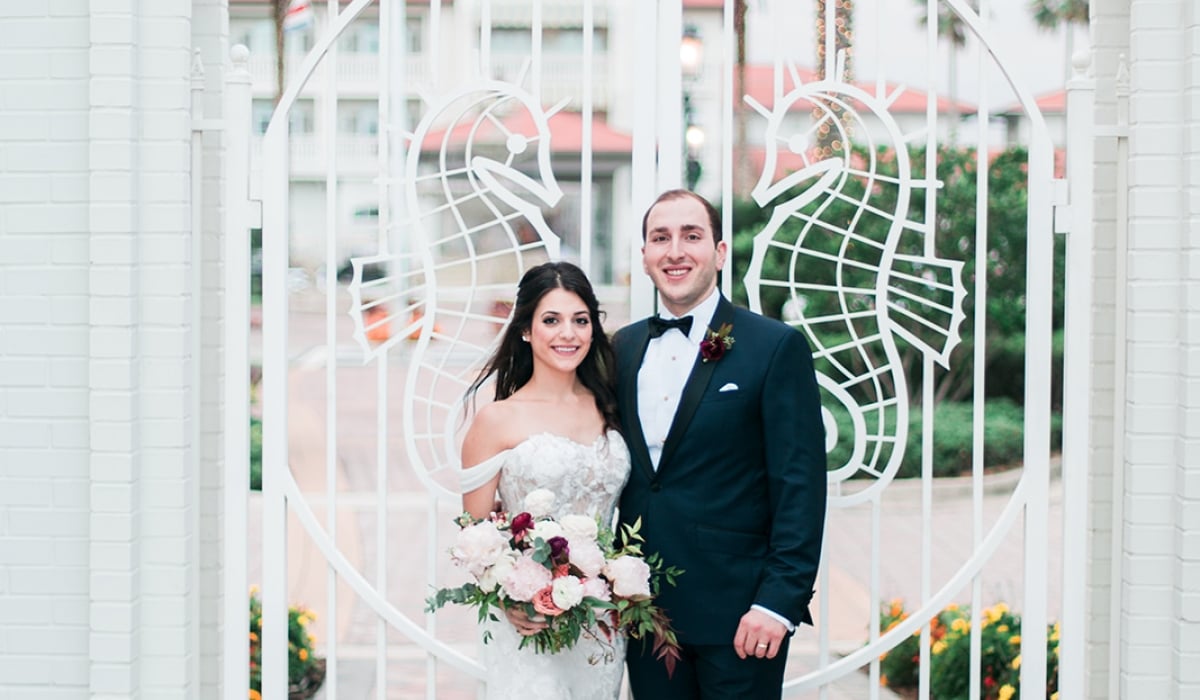 A bride and groom stand in front of a white gate decorated with seahorses, with Ponte Vedra Inn & Club in the background.
