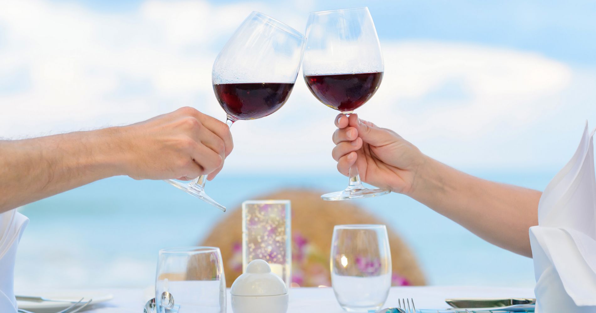 Two hands raise glasses of red wine over a table with the ocean in the background.